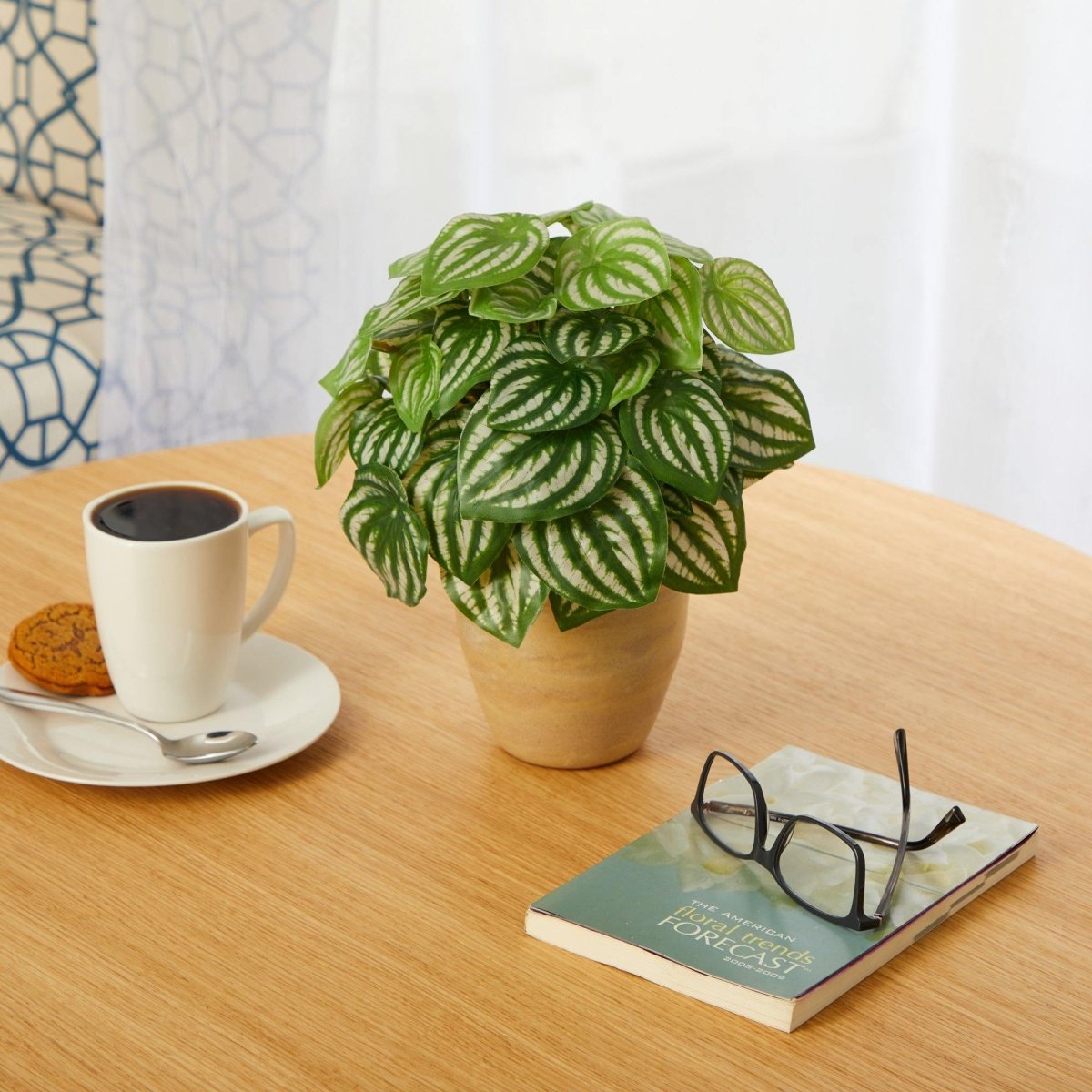 Potted artificial plant on a wooden table with a cup of coffee, book, and glasses.